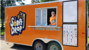 Soda Drop food trailer parked outside at an event with the owner looking out and smiling from the serving window.